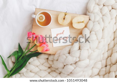Wooden tray with cup of tea, apple, spring tulips flowers, white merino wool plaid or blanket of thick yarn, card with text GOOD MORNING on white bedding. Breakfast in bed. Top view.