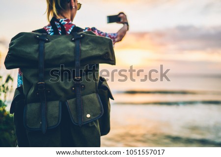 Selective focus on big green useful rucksack for traveling.Back view of hipster girl making photo on smartphone camera for share impressions.Cropped image of woman making picture of nature at sunset