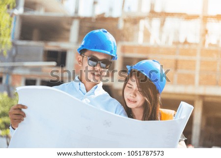 Asian man and woman engineer with the blue safety helmet meeting at the construction site with blueprints.
