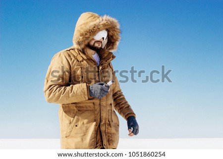 a guy in a brown jacket is standing in the winter on the street in the snow with a vape in his hands and looks into the distance