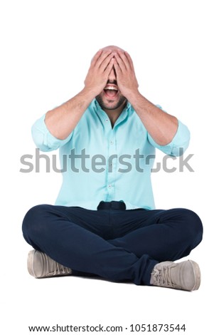 A young man sitting on the floor crossed legs covering his eyes and screaming with an open mouth, isolated on a white background.