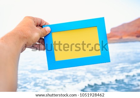 A male hand holds a blue photo frame with empty space seascape on background. Travel concept.