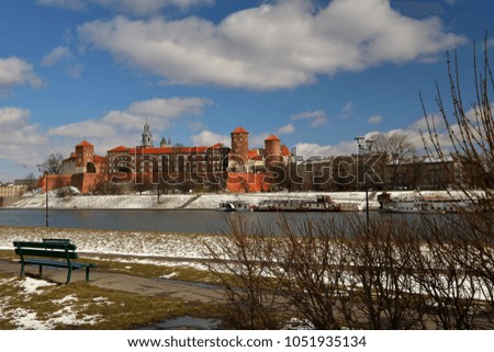 Krakow, Poland, view at small bushes and Vistula river's bank in foreground, with green bench, in background castle on hill, blue sky with picturesque white clouds, winter sunny day