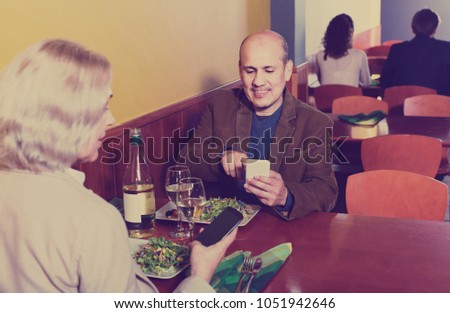 Positive senior couple busy with phones on date in cafe 