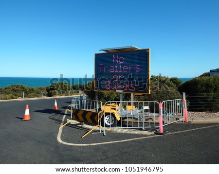 A three row fixed character matrix LED sign warns "No trailers - or buses " placed on a barricade in a beach car park