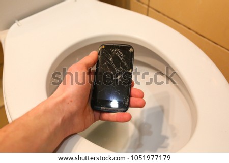 Man is holding a crashed black smartphone in hand over the toilet bowl. Broken lcd touch screen in the bathroom. Information technology accident photo. Guy is dropping phone in the toilet.