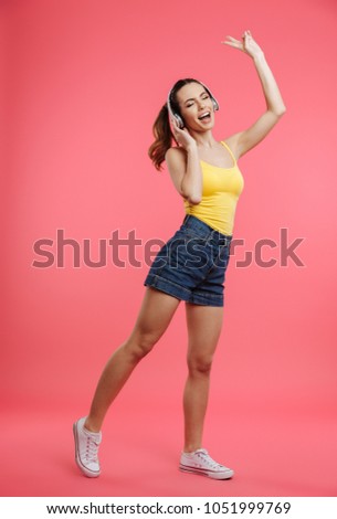 Full length portrait of a cheery young girl dressed in summer clothes listening to music with headphones and dancing isolated over pink background
