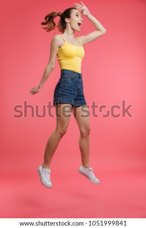 Full length portrait of a happy young girl dressed in summer clothes looking far away while jumping isolated over pink background