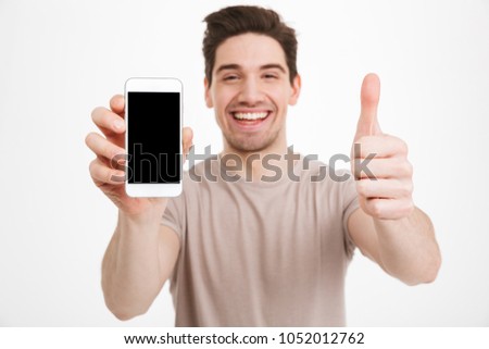 Photo of man in beige t-shirt demonstrating copyspace black screen of smartphone and gesturing thumb up isolated over white wall