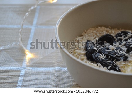Bowl of oatmeal porridge with peanuts, prunes, coconut flakes and butter on teal rustic table, hot and healthy food for Breakfast. Light tablecloth in a cage or plaid. Lights or garland on background.