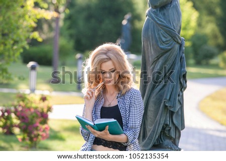 Blonde pretty woman posing with school books in park in summer. Young blond girl smiling with a book in the park on the campus of the university. Happy Female Student In Public Park. 