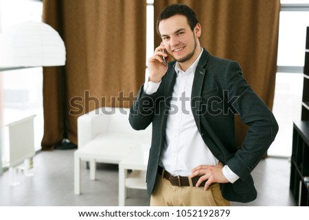 The young young man happily talks on the phone in the office against the background of brown curtains