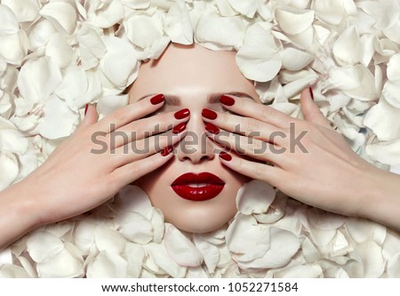 Portrait of a beautiful young girl in white rose petals. Red lips, red fingernails. The girl closes her eyes with her hands.Fashion, beauty, make-up, cosmetics, beauty salon, style, personal care.