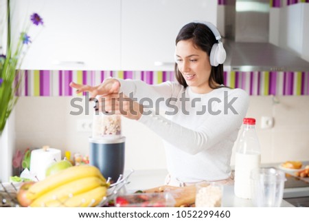 Young bpretty happy girl blending smoothie mixture while listening to music on headphones and standing in front of the kitchen counter.