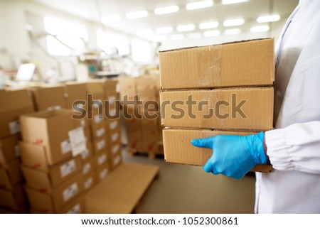 Close up view of a worker carrying a stack of packages in brown boxes within production industry storage room while wearing sterile cloths and rubber gloves.