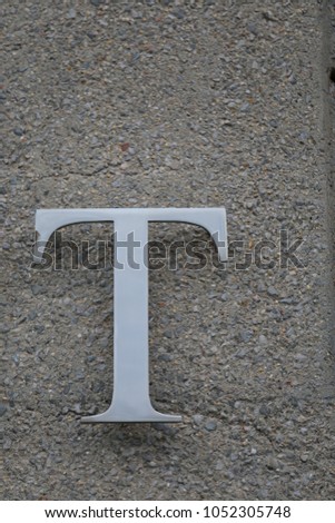 Close up outdoor view of the letter T fixed on a grey rough wall. Metallic isolated element in 3d. Capital letter of the alphabet. Abstract design. Sign visible in a french street.    