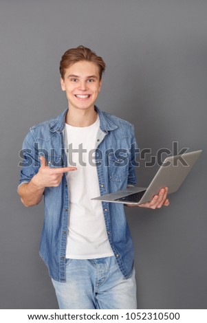 Young man in jeans jacket studio isolated on grey pointing at laptop screen cheerful