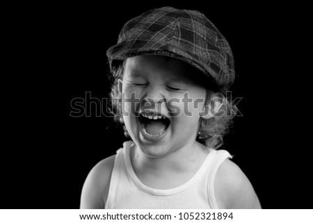 A portrait of a happy little boy laughing and smiling. He is wearing a white tank top and a plaid hat. The black and white headshot is of a child model and a child actor. The boy is playful, smiley. 