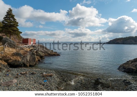 A small red building sits on the edge of a cliff in a small cove. The building has a wooden walkway leading to a beach, The ocean is smooth and calm on a bright sunny day with white fluffy clouds. 