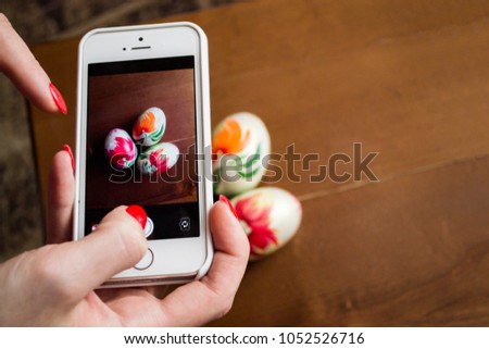 Woman taking picture of three handmade easter eggs on wooden table with her smartphone. Top view