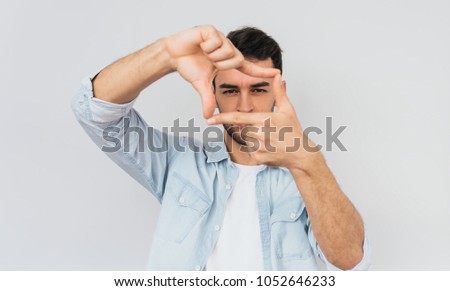 Studio portrait of handsome bearded male is looking through a frame formed by his hands. Serious expression of the face, isolated on a white background with copy space for text. People concept