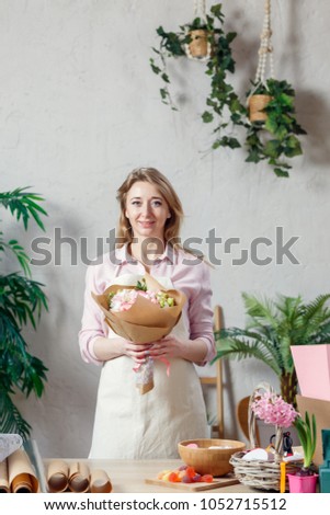 Photo of florist in apron with bouquet of flowers with kraft paper on background of indoor plants