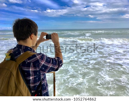 Close up young male hands make a photo at black smartphone. Bright and colorful picture of a man in shirt with backpack. Big and powerful sea waves on background beautiful clouds sky ocean.