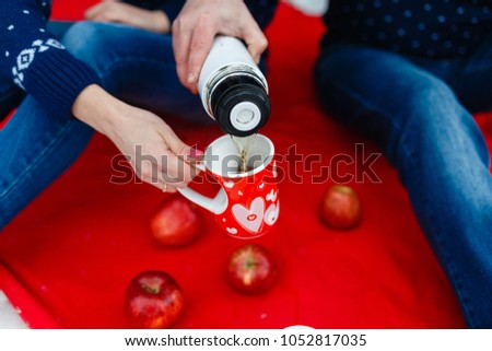 Man pours tea in a mug with a picture of the heart. Young couples in sweaters sit on red blanket. Wedding picnic in the fresh air in the winter in the forest or mountains. Offer to marry on a picnic.