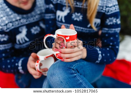 Mug of hot tea with a picture of a heart in the hands of a young couple in warm sweaters sitting on a red blanket. Wedding trip picnic outdoors in the winter in the woods or mountains.