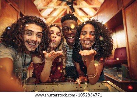 Group of friends enjoying a party and blowing confetti. Men and women capturing the fun in selfie at nightclub.