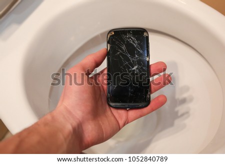 Man is holding a crashed black smartphone in hand over the toilet bowl. Broken lcd touch screen in the bathroom. Information technology accident photo. Guy is dropping phone in the toilet.