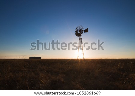 A broken windpump and a lone farm dam stand brave in a dry meadow of golden grass, while the sun sets against a blue cloudless sky. Royalty-Free Stock Photo #1052861669