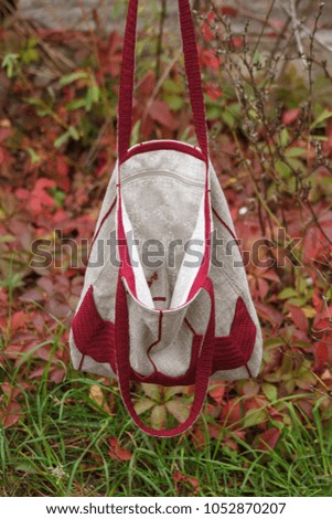 Crossbody bag is made of linen and decorated with knitted burgundy decor on the background of autumn leaves.