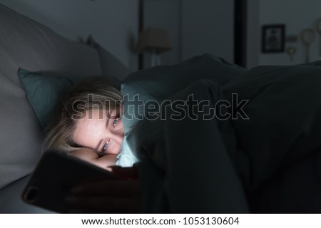 Portrait of young sleepy tired woman lying in bed under the blanket using smartphone at late night, can not sleep/ Insomnia, nomophobia, sleep disorder concept/ Dependency on a cell phone Royalty-Free Stock Photo #1053130604