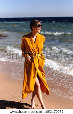Girl in a beautiful dress is walking along the beach on the seaside. The photo was made near the sea in a sunny warm day. The girl on the photo is wearing yellow long dress and big sunglasses.
