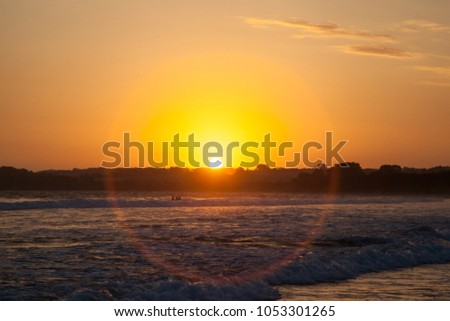 Sunburst on the horizon at sunset over the beach with waves and people swimming.