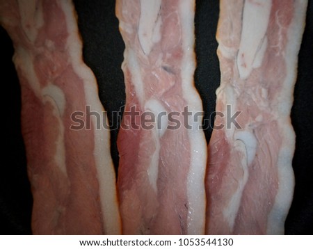 slices of raw bacon on black surface pan fried, close up