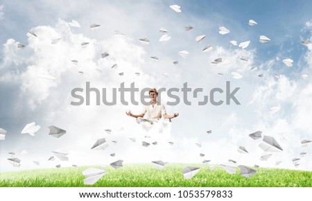 Young man keeping eyes closed and looking concentrated while meditating on cloud among flying paper planes with bright and beautiful landscape on background.