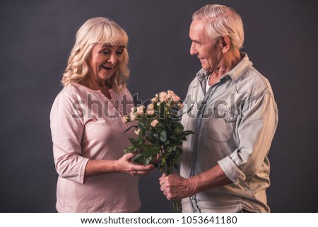 Beautiful old man and woman in casual clothes on gray background. Man is presenting flowers to his woman
