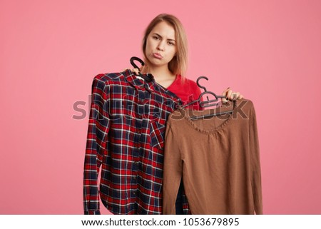Unhappy discontent female curves lips as holds two hangers with clothes, being upset as has not enough money for paying for purchase, isolated over pink background. People, shopping, problems
