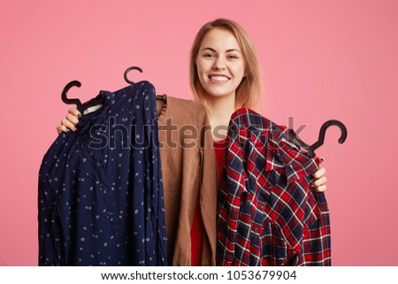 Happy lovely young woman holds clothes on hangers, rejoices new purchase, doesn`t know what to choose, going to spend money on new outfit, isolated over pink studio background. Shopping concept
