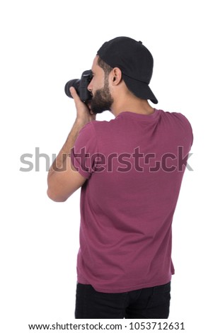 A rear shot of a young man standing, wearing a cap and T-shirt holding his camera taking photos as a professional photographer, isolated on a white background.
