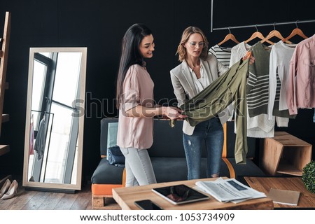 Nice color. Beautiful young woman helping to choose clothes to her customer while working in the fashion boutique Royalty-Free Stock Photo #1053734909