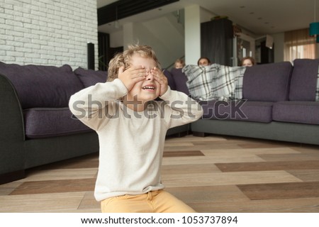 Kid boy playing hide and seek game at home, child closing eyes with hands counting while parents and sister hide behind sofa in living room peeking out, happy family having fun with children concept Royalty-Free Stock Photo #1053737894
