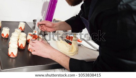 Faceless action shot of a french pastry chef filling cristal jars with a spoon and a pastry bag on a kitchen work space environment. Haute cuisine concept.