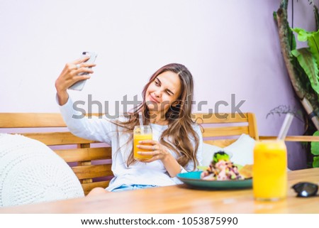 A beautiful woman in a cafe makes selfie on a smartphone, drinks freshly squeezed juice.
