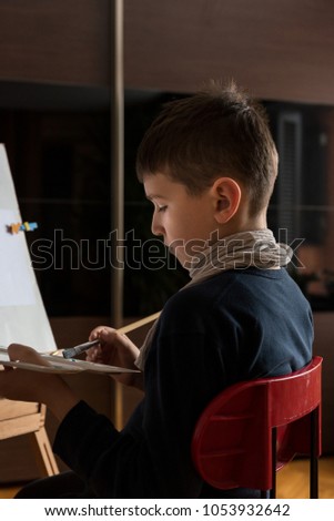 Young boy sitting in front of easel, painting with brush in his hand