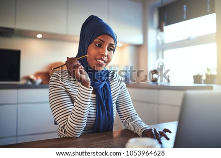 Young Arabic female entrepreneur sitting at a table in her kitchen writing down notes and working on a laptop