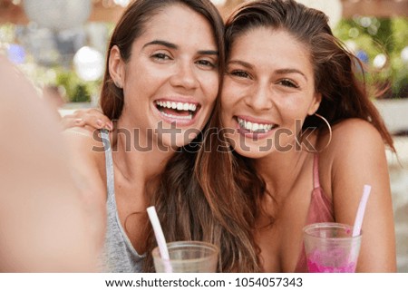 Glad European female sisters with broad smile pose at camera with happy expression, spend free time in terrace cafe or bar, drink fresh cocktails, pose for making selfie and sharing photos in internet