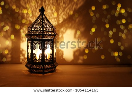 Ornamental Arabic lantern with burning candle glowing at night and glittering golden bokeh lights. Festive greeting card, invitation for Muslim holy month Ramadan Kareem. Dark background. Royalty-Free Stock Photo #1054114163